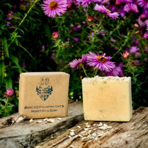 2 Anne's Exfoliating Oat & Honey Milk Soap one wrapped in brown paper one unwrapped on some wood with purple daisy type flowers behind them