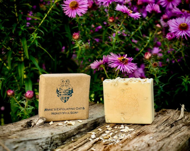 2 Anne's Exfoliating Oat & Honey Milk Soap one wrapped in brown paper one unwrapped on some wood with purple daisy type flowers behind them