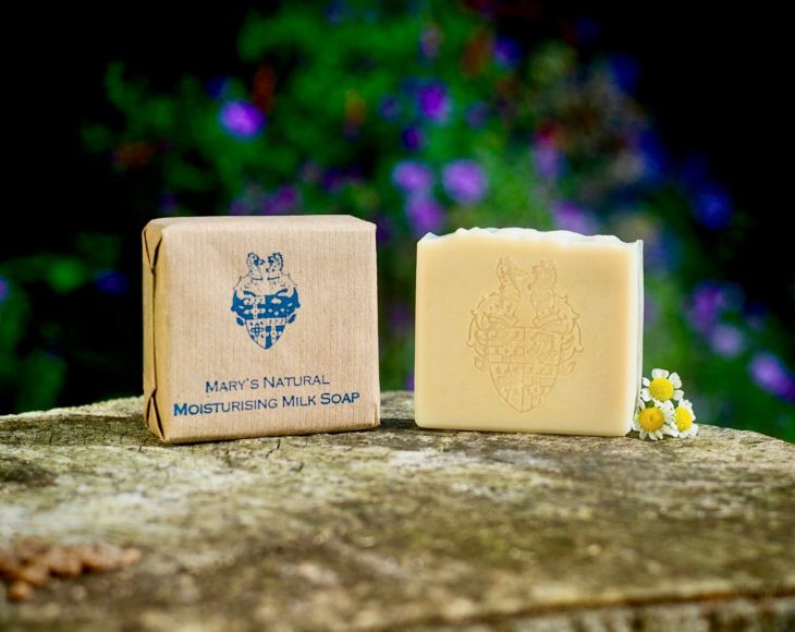 2 Mary's Natural Moistursing Milk Soaps one is wrapped in brown paper one is unwrapped on a slab of stone with 3 chamomile flowers next to and blurry purple flowers in the background