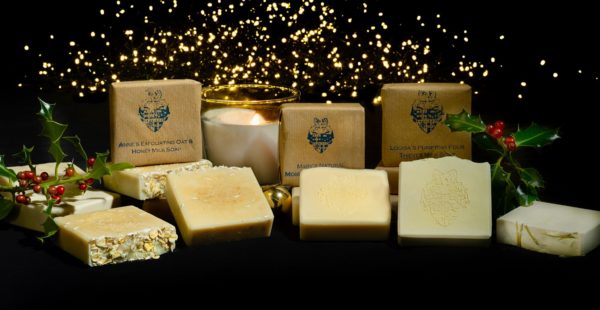 A selection of handmade soaps on a black cloth with some sprigs of holly and a candle and gold twinkly lights in the background